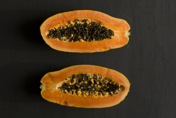 Две половинки папайи, разрезанные пополам, лежат на черном современном фоне. Черные семена сладкой апельсиновой папайи. Вегетарианство, здоровое питание, диета. Тропические фрукты. Вид сверху. Принято. Плоский лежал
. - Фото, изображение