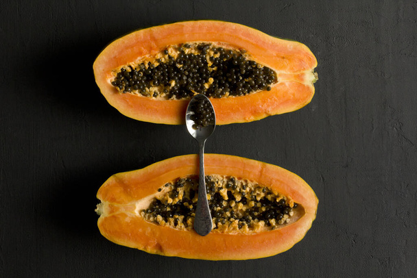 Two halves of papaya fruit cut in half, spoon, lie on black modern background. Black seeds of sweet, orange papaya. Vegetarianism, healthy nutrition. Tropical fruits.Top view. Copy space. Flat lay. - Photo, Image