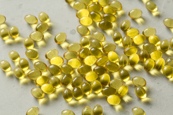 Pile of transparent capsules of golden yellow color lies on light gray modern background. Oil filled capsules vitamin A, vitamin D3, fish oil, omega 3, 6, 9, evening primrose, vitamin D, vitamin E. - 写真・画像