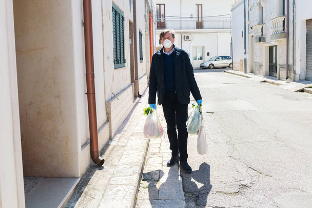 Pusta ulica, mężczyzna wraca do domu z zakupami z zakupów w ochronnych maskach i rękawiczkach, aby powstrzymać rozprzestrzenianie się koronawirusu we Włoszech podczas kwarantanny. Przestrzeganie norm zdrowotnych covid-19 - Zdjęcie, obraz