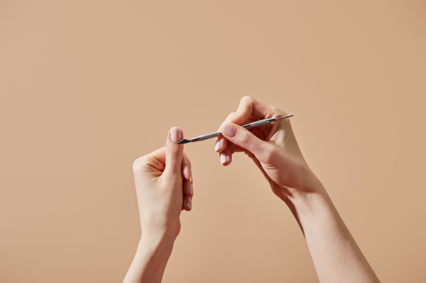 Vista cortada da mulher fazendo manicure usando empurrador de cutícula isolado no bege
 - Foto, Imagem