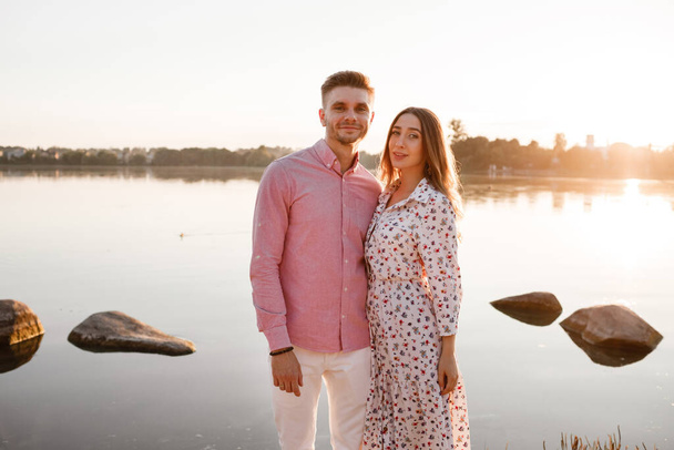 Liebespaar, das sich bei Sonnenuntergang am See umarmt. Schönes junges verliebtes Paar, das bei Sonnenuntergang im hellen Licht am Ufer des Sees spaziert. Kopierraum - Foto, Bild