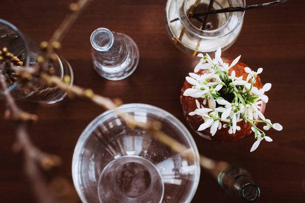 Gotas de neve em um vaso de barro entre recipientes de vidro na mesa. Conceito para a primavera. As primeiras flores. Ainda vida
 - Foto, Imagem