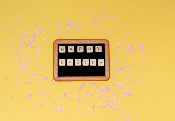 Chalk board with the words Happy Easter and pink decorative paper confetti on a yellow background, copy space, flat lay. The concept of the holiday, spring. View from above. - Фото, зображення