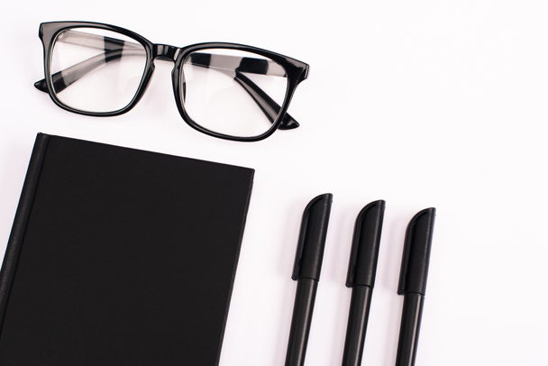 κορυφή άποψη των στυλό κοντά γυαλιά και μαύρο σημειωματάριο που απομονώνονται σε λευκό  - Φωτογραφία, εικόνα