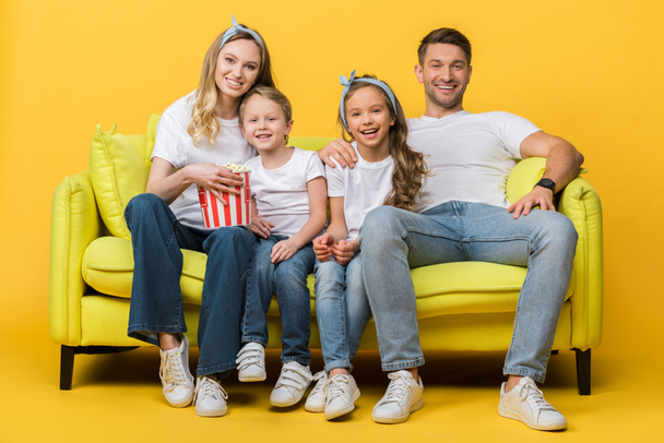 smiling parents and kids watching movie on sofa with popcorn bucket on yellow  - Zdjęcie, obraz