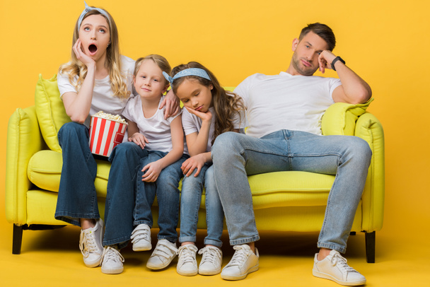 bored parents and kids watching movie on sofa with popcorn bucket on yellow  - Φωτογραφία, εικόνα