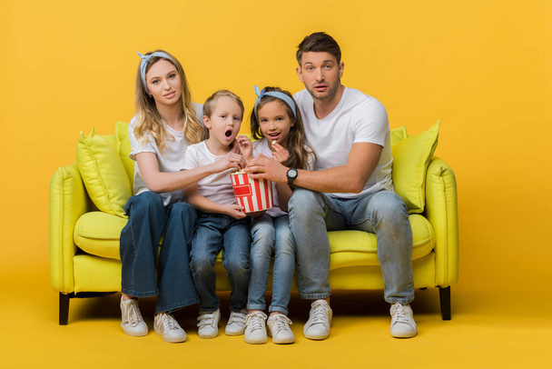 surprised family watching movie on sofa with popcorn bucket on yellow  - Φωτογραφία, εικόνα