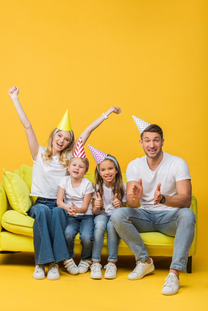 happy family in birthday party caps applauding and showing thumbs up on sofa on yellow - Zdjęcie, obraz
