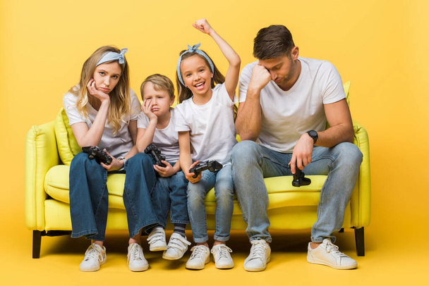 KYIV, UKRAINE - Março 4, 2020: família chateada com joysticks sentados no sofá com a filha feliz ganhar no jogo de vídeo em amarelo
 - Foto, Imagem