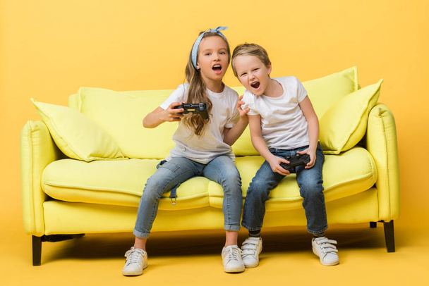 KYIV, UKRAINE - MARCH 4, 2020: emotional siblings screaming and playing video game with joysticks while sitting on sofa on yellow - Фото, изображение