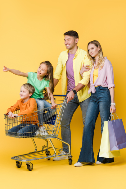 parents with shopping bags and kids gesturing and sitting in shopping cart on yellow  - Foto, Imagen
