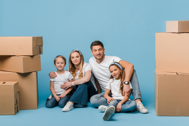 happy parents hugging kids and sitting on blue with cardboard boxes for relocation - Foto, Bild