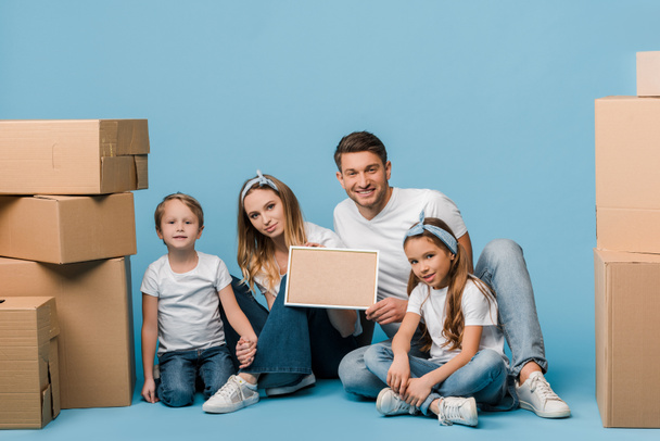 happy parents and kids holding frame and sitting on blue with cardboard boxes for relocation - Foto, Imagem
