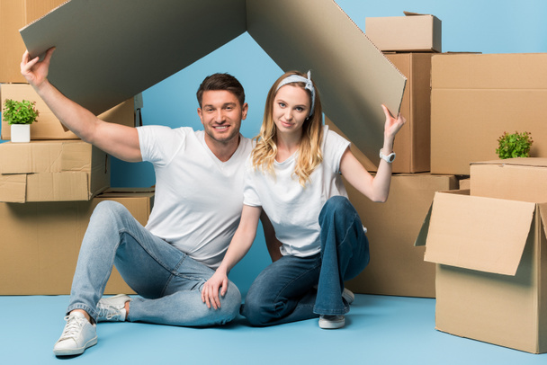 beautiful couple holding carton roof over heads while sitting on blue with cardboard boxes for relocation - Photo, Image