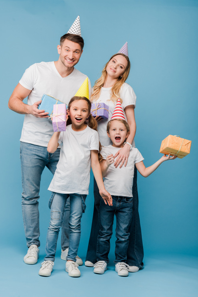 excited parents and kids in birthday party caps holding gift boxes on blue - Foto, Imagen
