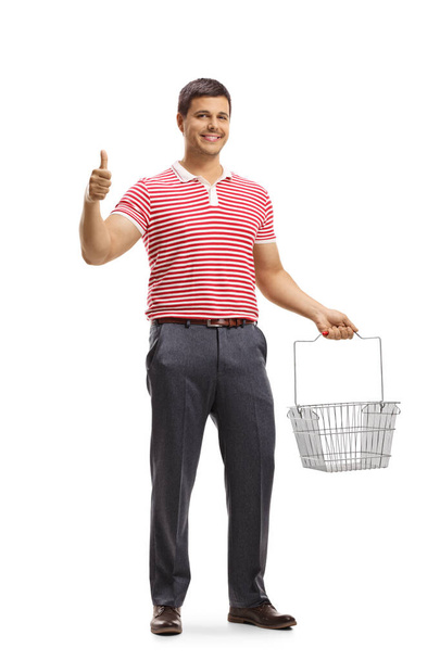 Full length portrait of a young man holding an empty shopping basket and gesturing thumb up sign isolated on white background - Фото, изображение