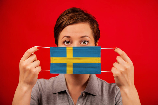 Coronavirus COVID-19 in Sweden. Woman in medical protective mask with the image of the flag of Sweden. The concept of preventing the spread of the epidemic and treating coronavirus. - Фото, изображение