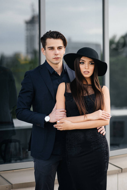 A beautiful, stylish pair of young people in black clothes and glasses stand against the background of an office building in the sunset. Fashion and style - Фото, изображение