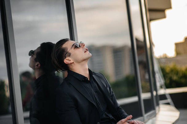 Un joven con estilo se encuentra cerca de un edificio de espejos durante la puesta del sol. Moda - Foto, Imagen