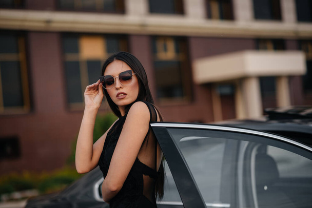 Stylish young girl stands near the car in a black dress. Business fashion and style - Foto, immagini