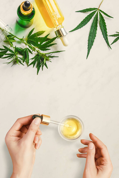 Pipet met CBD cosmetische olie in vrouwelijke handen op een tafel achtergrond met verschillende flessen met cannabis olie en hennepbladeren, marihuana - Foto, afbeelding