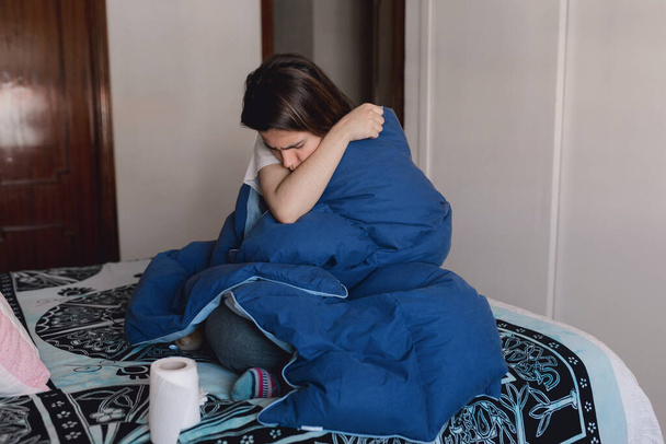 Jovem mulher doente infectada pelo coronavírus está tossindo em sua manga superior enquanto cobre com uma colcha azul em sua casa
 - Foto, Imagem