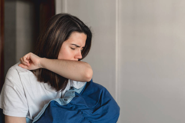 Mladá nemocná žena infikovaná koronavirem kašle do horního rukávu, zatímco ve svém domě přikrývá modrou dekou - Fotografie, Obrázek