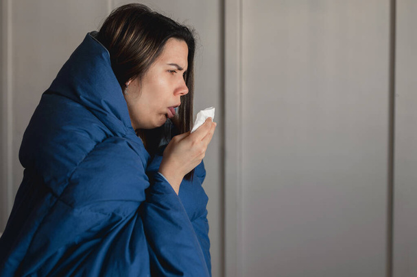 Молода хвора жінка, заражена коронавірусом, кашляє в тканину, в той час як вона вкрита блакитним ковдрою в безпеці свого дому. Епідемія вірусу в Іспанії. - Фото, зображення