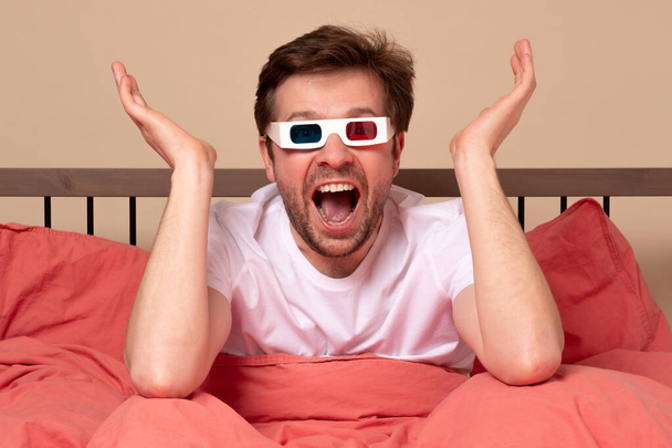 Fiatal kaukázusi férfi otthon tévézik az ágyban 3D-s szemüvegben, izgatottan és ámulatba ejtve. Stúdió felvétel - Fotó, kép