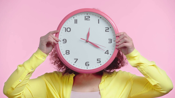 fröhliches Mädchen lächelt und verschleiert Gesicht mit runder Uhr isoliert auf rosa - Filmmaterial, Video