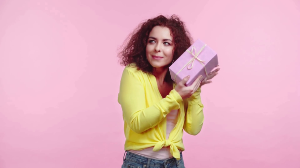 περίεργο σγουρό κορίτσι κουνώντας κουτί δώρου απομονωμένο σε ροζ - Πλάνα, βίντεο