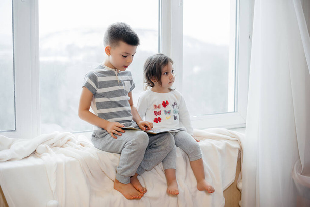 Brother and sister are sitting on the windowsill and reading a book. Happiness, family - Fotoğraf, Görsel