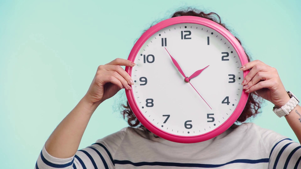 cheerful curly girl obscuring face with round clock isolated on turquoise - Footage, Video