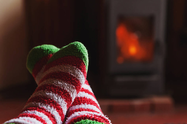 Πόδια σε χειμερινά ρούχα μάλλινες κάλτσες στο φόντο τζάκι. Γυναίκα κάθεται στο σπίτι το χειμώνα ή το φθινόπωρο βράδυ χαλάρωσης και προθέρμανσης. Χειμώνας και κρύο έννοια του καιρού. Παραμονή Χριστουγέννων Hygge - Φωτογραφία, εικόνα