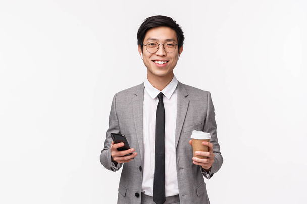 Irodai dolgozók, üzleti és életmód koncepció. Derékig érő portré sikeres fiatal ázsiai üzletemberről öltönyben, vezeték nélküli fejhallgatóval, kezében okostelefonnal és elvitelre szánt kávéval, mosolygós fényképezőgéppel - Fotó, kép