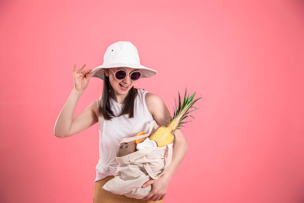 Portrait of a young stylish woman dressed in summer clothes with a hat and sunglasses, holding an ECO-fruit bag, on a pink isolated background. - Photo, Image