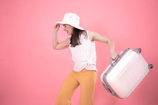 retrato de una mujer con estilo en ropa de verano de moda y un sombrero blanco sobre un fondo rosa con una maleta para viajar. Concepto de turismo de verano
. - Foto, imagen