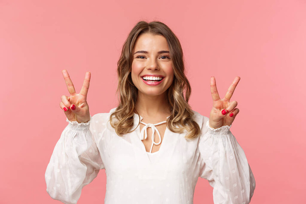 Nahaufnahme Porträt eines optimistischen kawaii jungen blonden Mädchens mit einem schönen strahlenden Lächeln, das Friedenszeichen zeigt und mit positiver Einstellung in die Kamera blickt, den Frühling genießt, weißes Kleid, rosa Hintergrund - Foto, Bild
