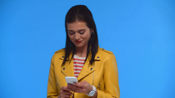 Χαμογελαστή γυναίκα που χρησιμοποιεί smartphone και δείχνει ναι χειρονομία που απομονώνεται στο μπλε - Πλάνα, βίντεο