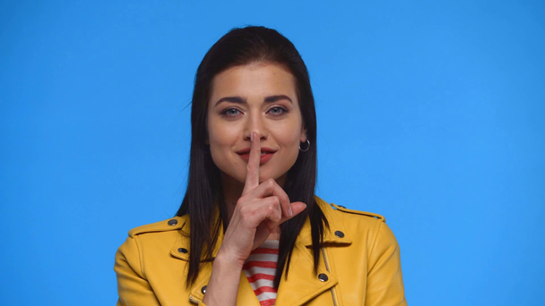 Atraktivní dívka ve žluté bundě ukazující mlčení znamení izolované na modré - Záběry, video