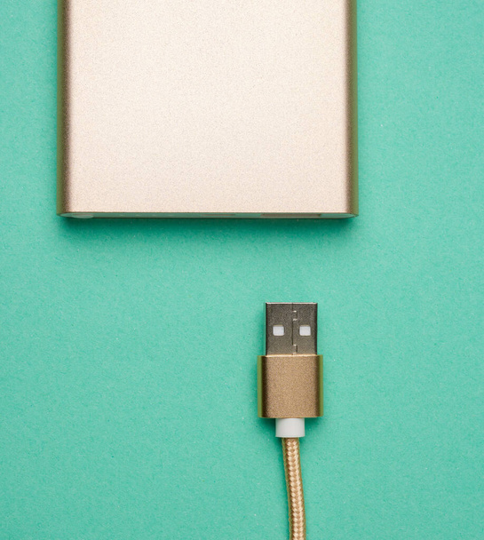 golden power bank and cord with a usb connector for recharging mobile devices on a green background, top view - Photo, Image