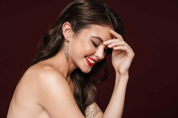Image of joyful shirtless woman wearing earrings laughing and looking downward isolated over burgundy background - Photo, image
