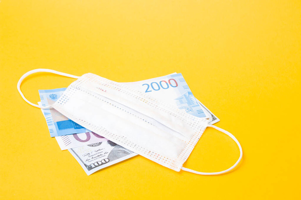 three banknotes from different countries, dollars, rubles and euros are under a protective medical mask of white color, yellow background, copy space, crisis 2020, pandemic and quarantine concept - Fotografie, Obrázek