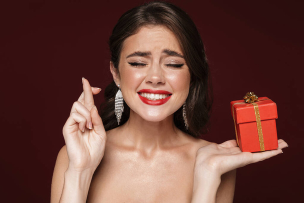 Image of nice shirtless woman wearing earrings with fingers crossed and holding gift box isolated over burgundy background - Φωτογραφία, εικόνα