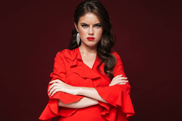 Image of displeased beautiful woman wearing red dress looking at camera with arms crossed isolated over burgundy background - Photo, Image