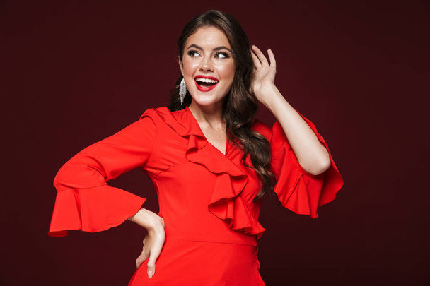 Image of excited woman wearing red dress posing and smiling isolated over burgundy background - Zdjęcie, obraz