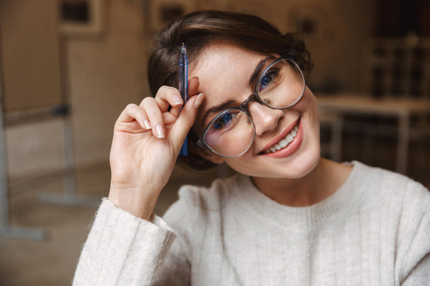 Imagem de jovem bela mulher caucasiana vestindo óculos sorrindo e segurando caneta no café
 - Foto, Imagem
