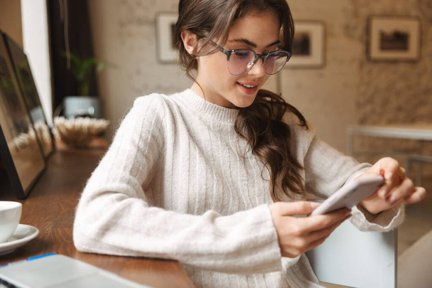 Image of young beautiful caucasian woman wearing eyeglasses using smartphone in cafe - Foto, afbeelding