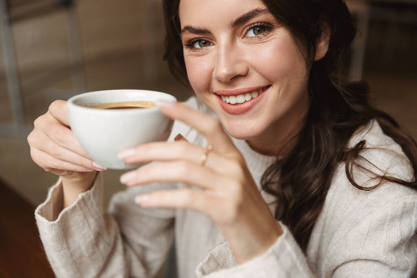 Изображение молодой красивой белой женщины с длинными каштановыми волосами, улыбающейся во время питья кофе в кафе
 - Фото, изображение